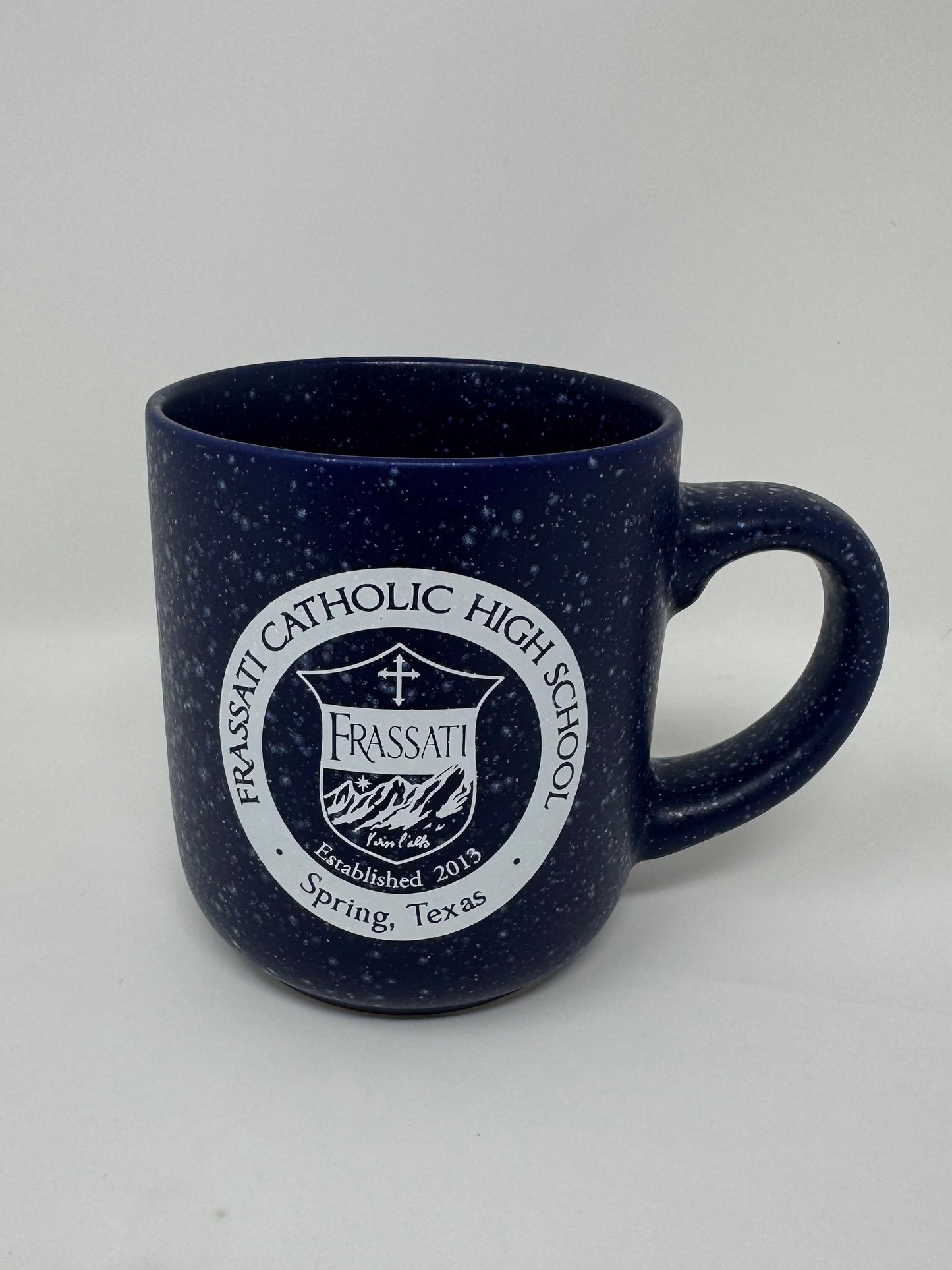 A dark navy blue speckled ceramic mug with a white printed circular logo for Frassati Catholic High School, shown on a plain light background.